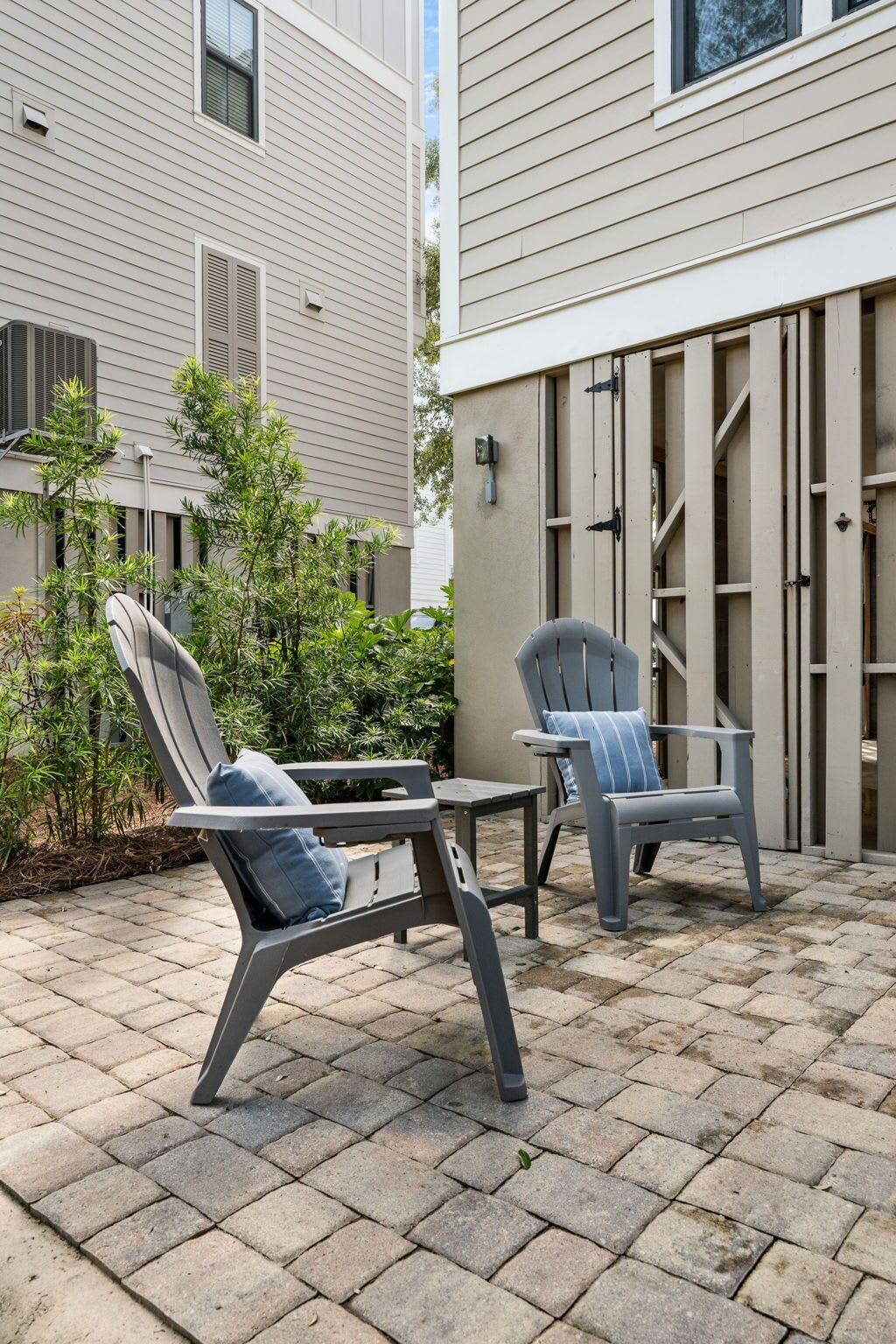 An outdoor patio with two grey lounge chairs and a small table, surrounded by a paved area and greenery.