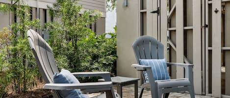 An outdoor patio with two grey lounge chairs and a small table, surrounded by a paved area and greenery.