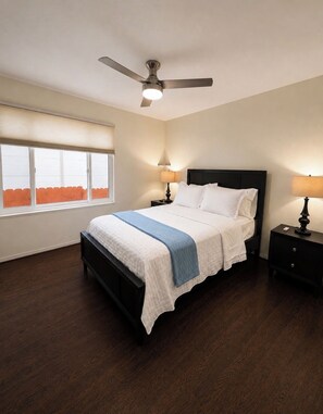A bedroom with a bed, a ceiling fan, two bedside tables with lamps, a window with blinds, and a view of a fence outside.