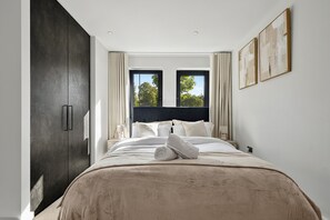 Un dormitorio moderno con una cama grande, un cabecero oscuro y dos ventanas con cortinas.