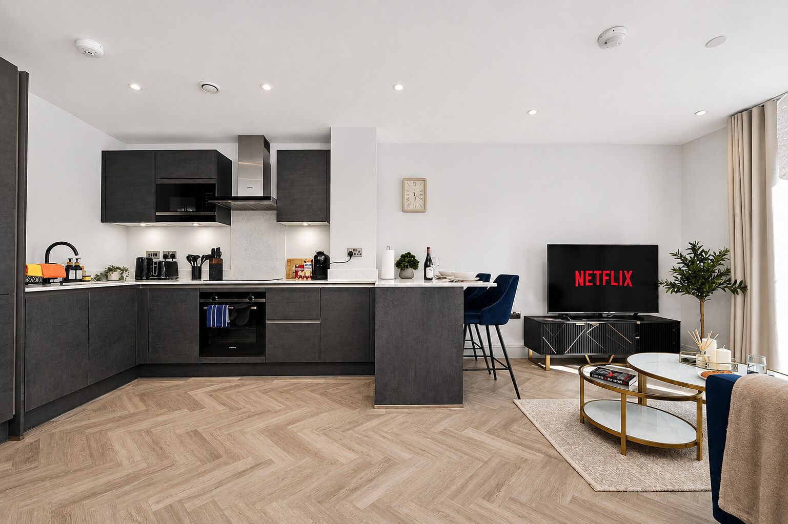 A modern kitchen with a built-in island, a dining area with a TV displaying 'NETFLIX', and a living space with a coffee table and a plant.