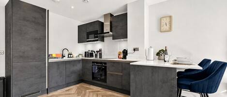 A modern kitchen with dark cabinetry, a central island, and a dining area with blue chairs.