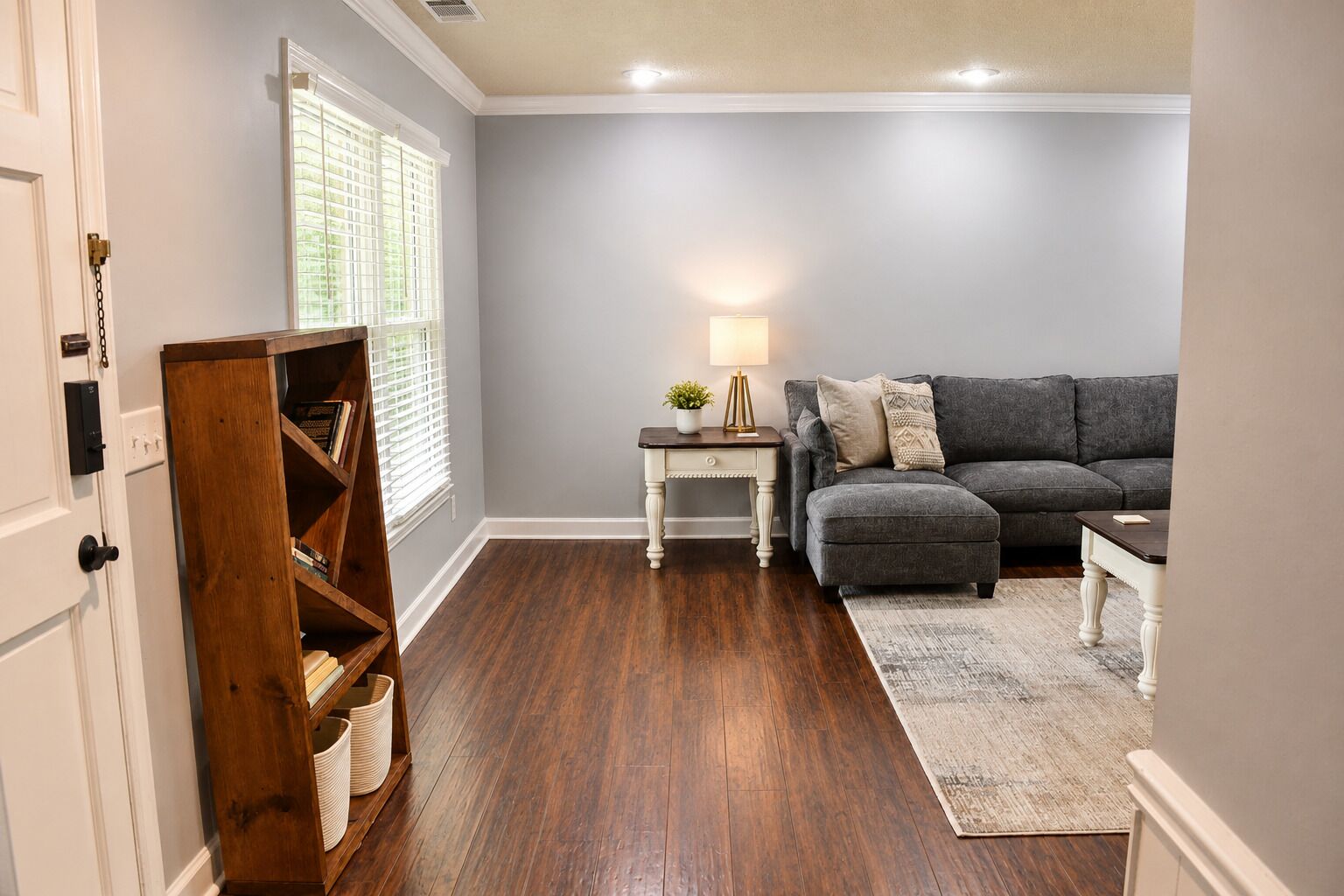 A living room with a grey sofa, a wooden bookshelf, a small table with a lamp, and a patterned rug.