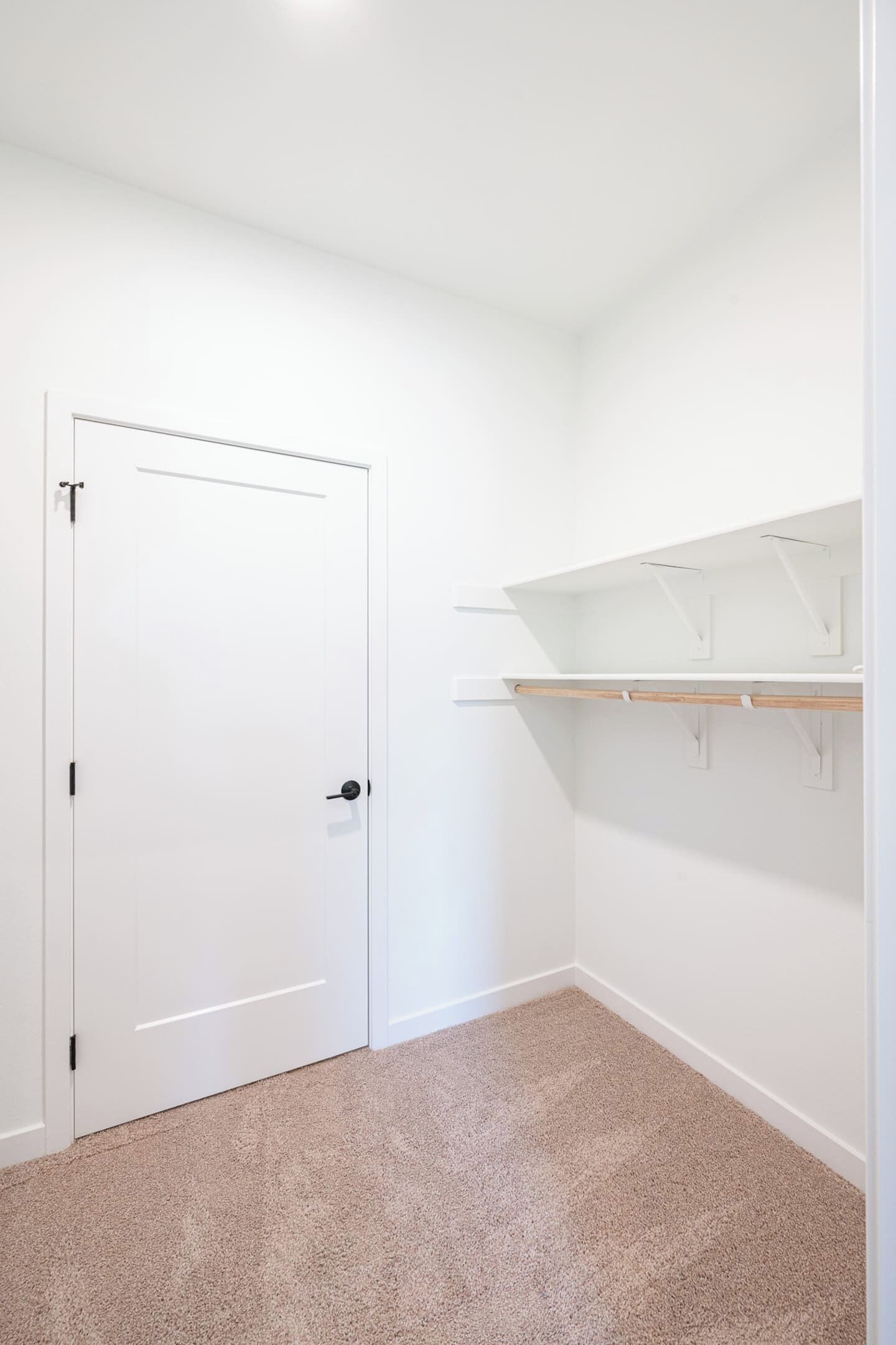 Ein Zimmer mit weißer Tür, beigem Teppich und weißen Wänden. An der Wand befinden sich zwei weiße Regale mit Haken.