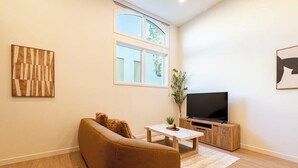 A living room with a sofa, a coffee table, a TV on a wooden stand, and a window with a view of greenery.