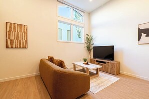 Un salon comprenant un canapé, une table basse, un téléviseur posé sur un meuble en bois et une fenêtre donnant sur un paysage verdoyant.