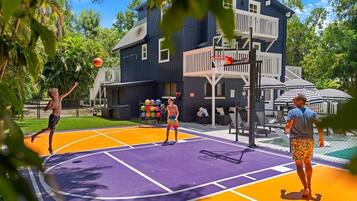 An outdoor basketball court with a purple and orange playing surface, surrounded by a fence, with a building in the background.