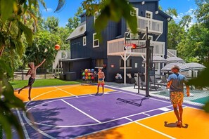 En utomhusbasketplan med ett lila och orange underlag, omgiven av ett staket, och en byggnad syns i bakgrunden.