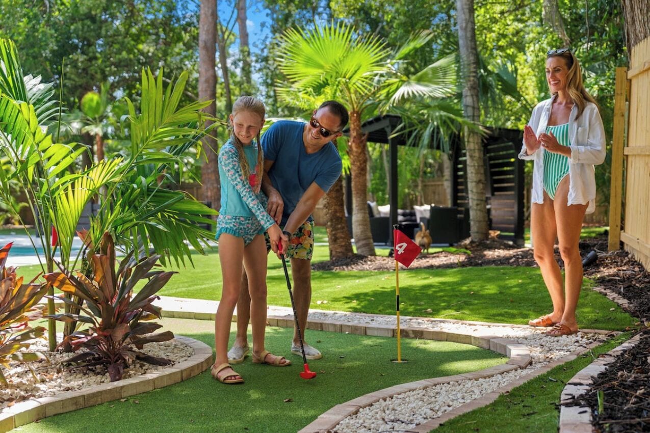 Une personne jouant au mini-golf dans un jardin agrémenté de plantes tropicales et d’une petite piscine.