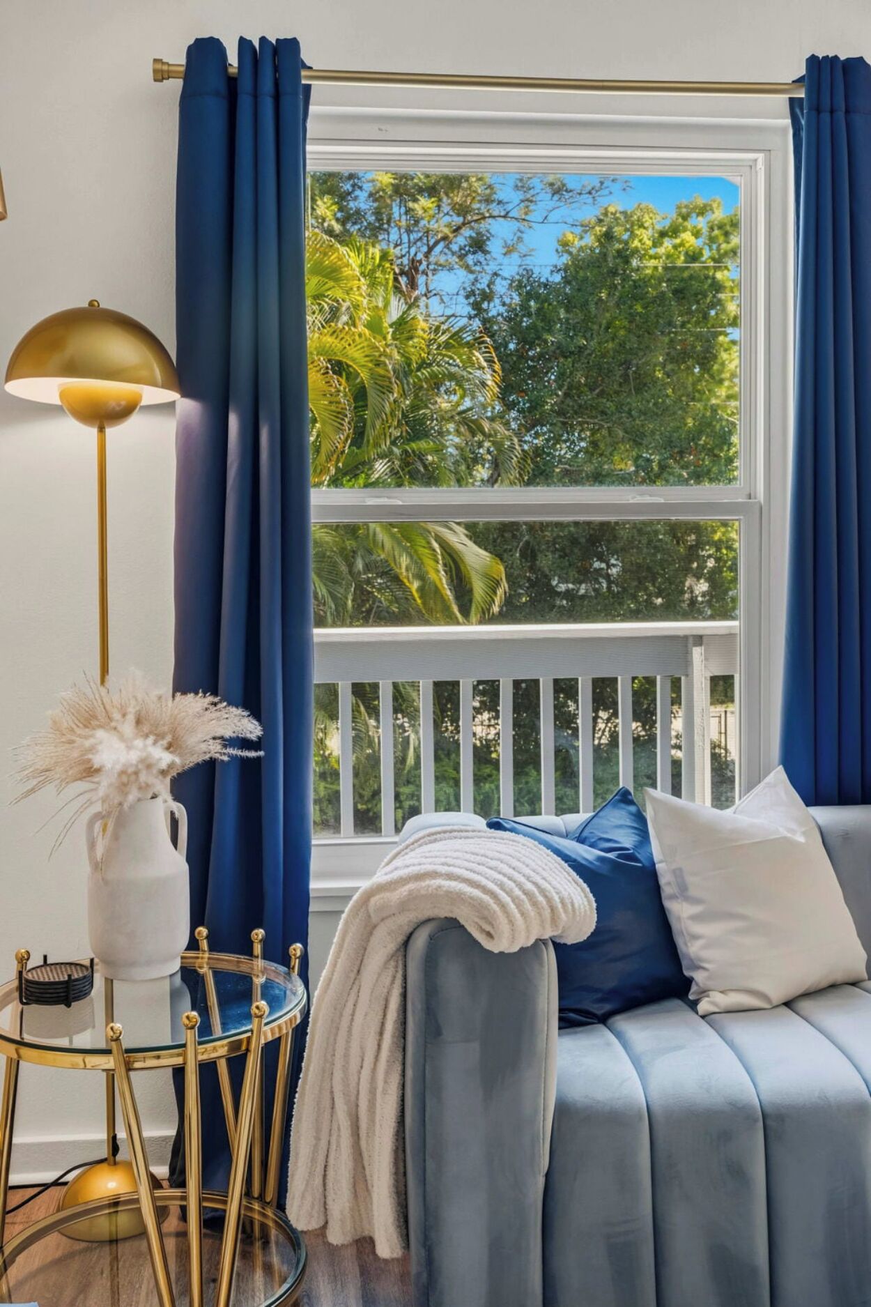 A room with a view of trees through a window, a sofa with pillows, a lamp, and a vase with dried flowers.