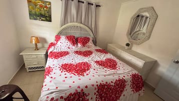 A bedroom with a bed covered in red heart-shaped decorations, a white headboard, a bedside table with a lamp, a dresser, and a mirror on the wall.