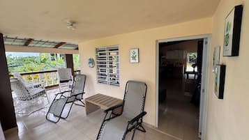 A covered outdoor seating area with two chairs and a table, a window with metal bars, and a view of a garden.