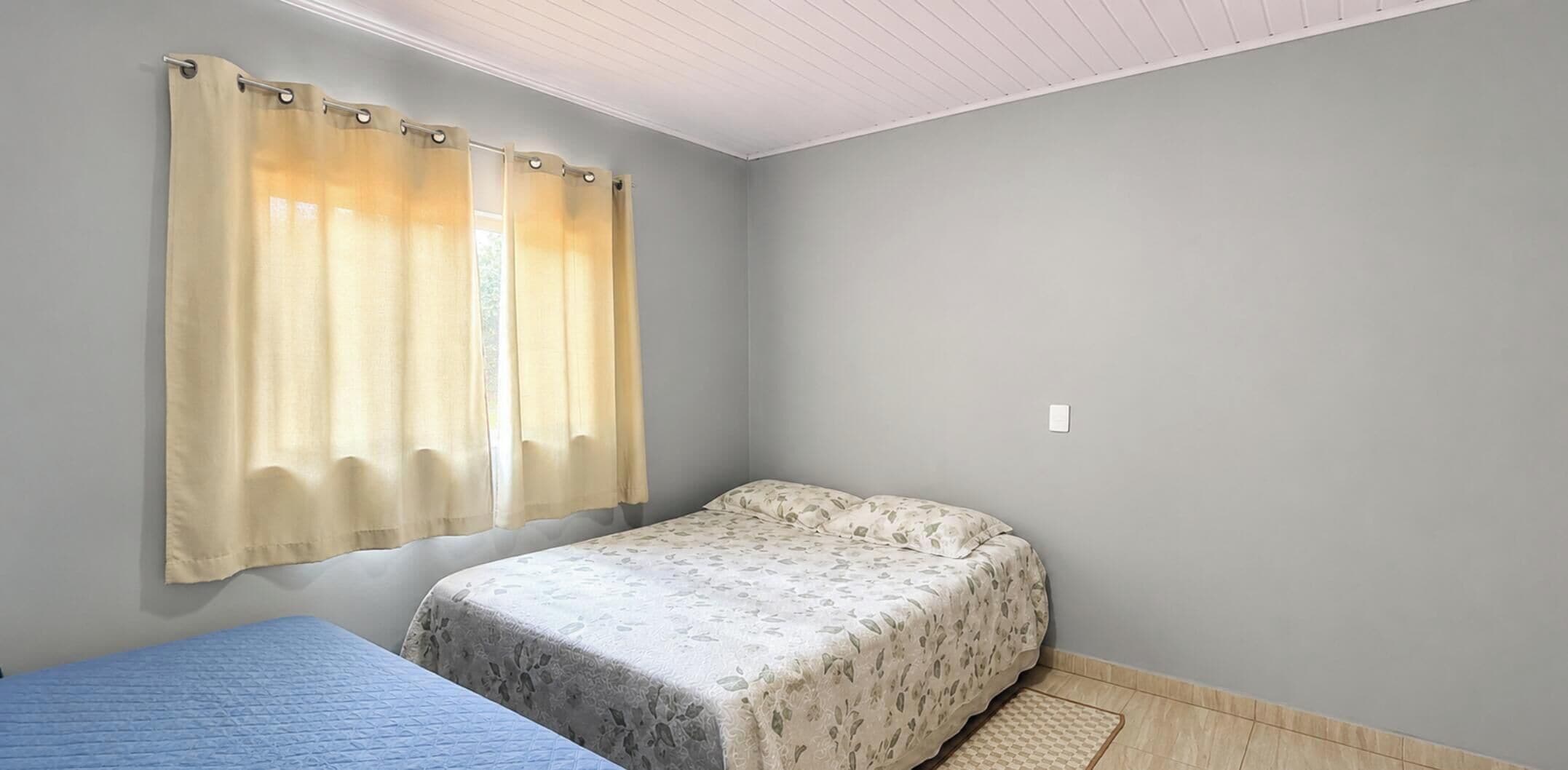 A room with a single bed, a window with curtains, a light switch on the wall, and a small rug on the floor.