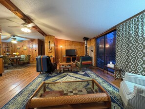A cozy living room with a fireplace, wooden walls, a television, and a large window. - Riverside Retreat in Morley, MI (Morley)