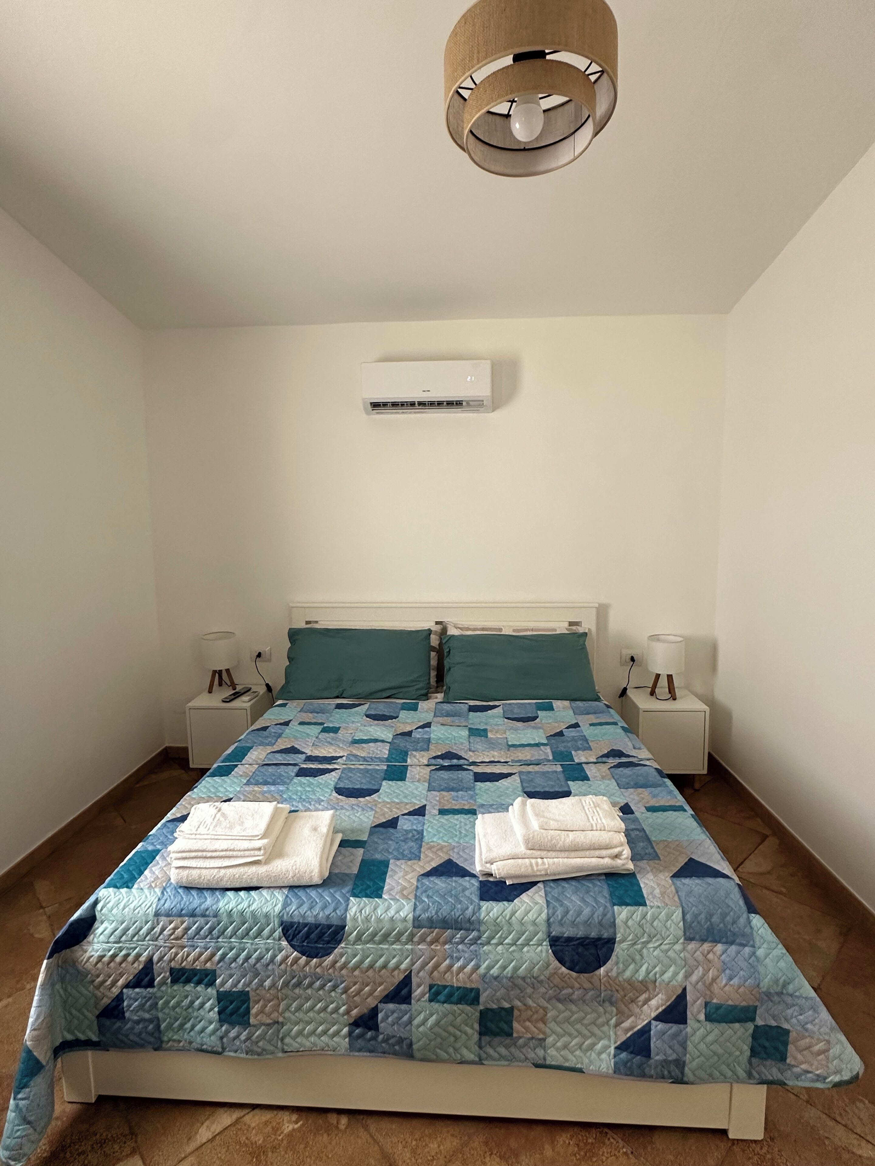 A bedroom with a bed, two bedside tables, a wall-mounted air conditioner, and a ceiling light fixture.