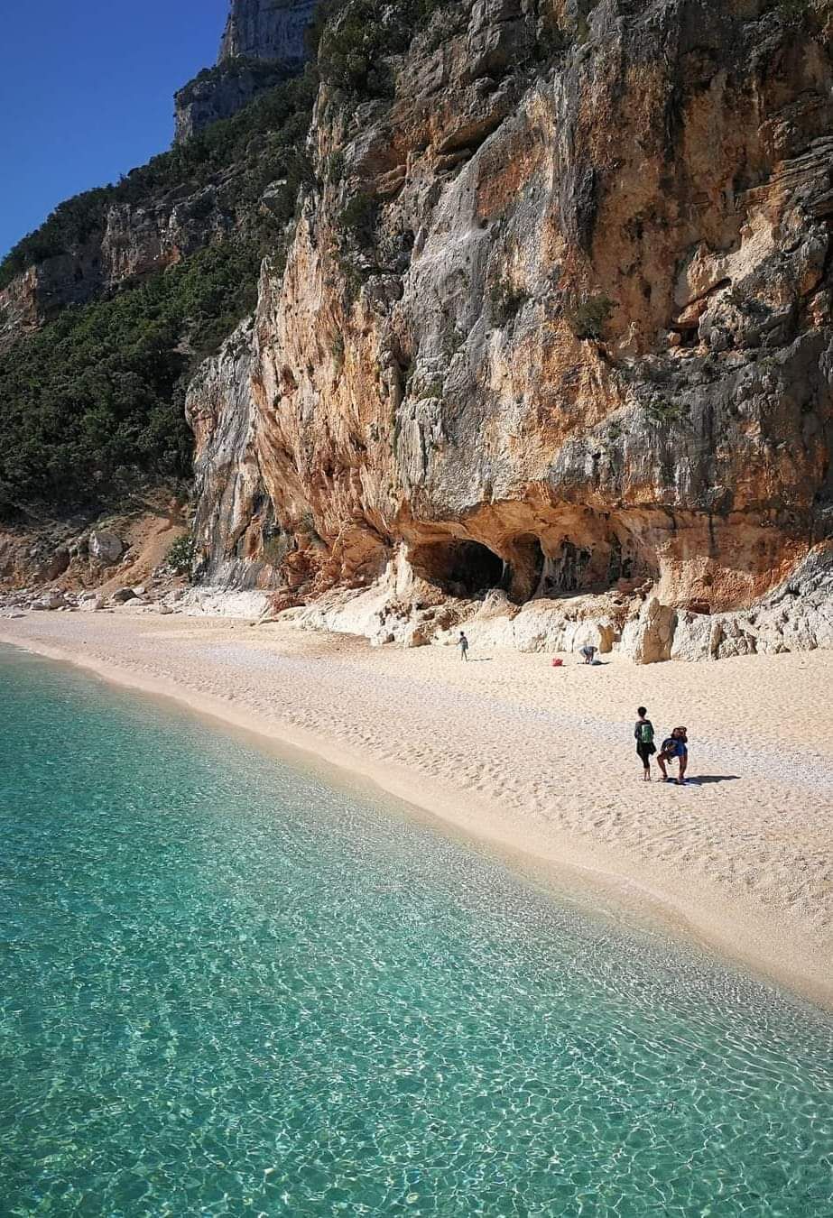 Ein Strand mit klarem türkisblauem Wasser, einer felsigen Klippe und einem Höhleneingang.