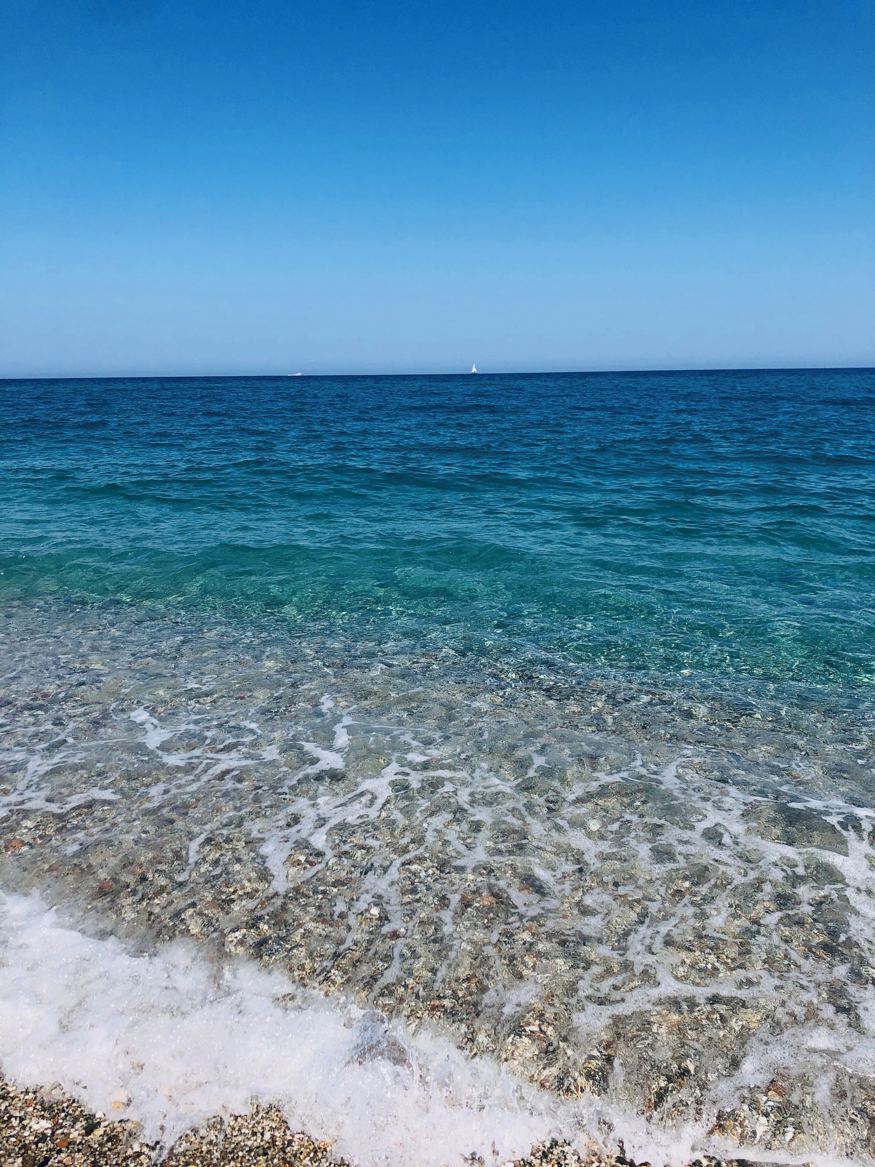 Freie Sicht auf einen Strand mit flachem Wasser, das den Meeresboden zeigt, ein entferntes Segelboot und ein klarer, blauer Himmel.