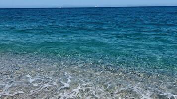 Freie Sicht auf einen Strand mit flachem Wasser, das den Meeresboden zeigt, ein entferntes Segelboot und ein klarer, blauer Himmel.