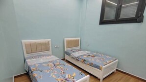 A room with two single beds, wooden flooring, a window with blinds, and a ceiling light fixture.