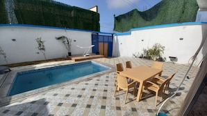 A swimming pool with tiled flooring, a table with chairs, and potted plants.