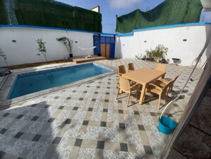 A swimming pool with tiled flooring, a table with chairs, and potted plants.