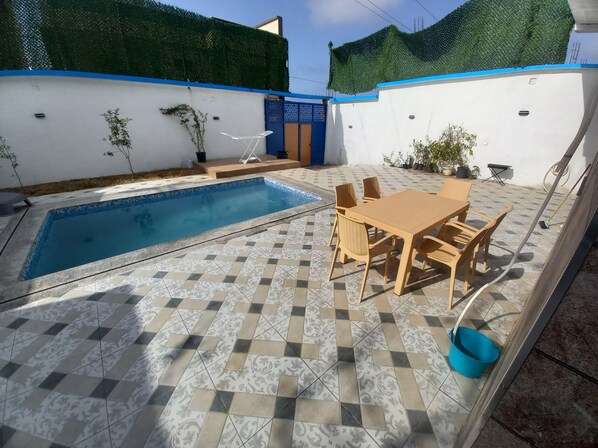 A swimming pool with tiled flooring, a table with chairs, and potted plants.