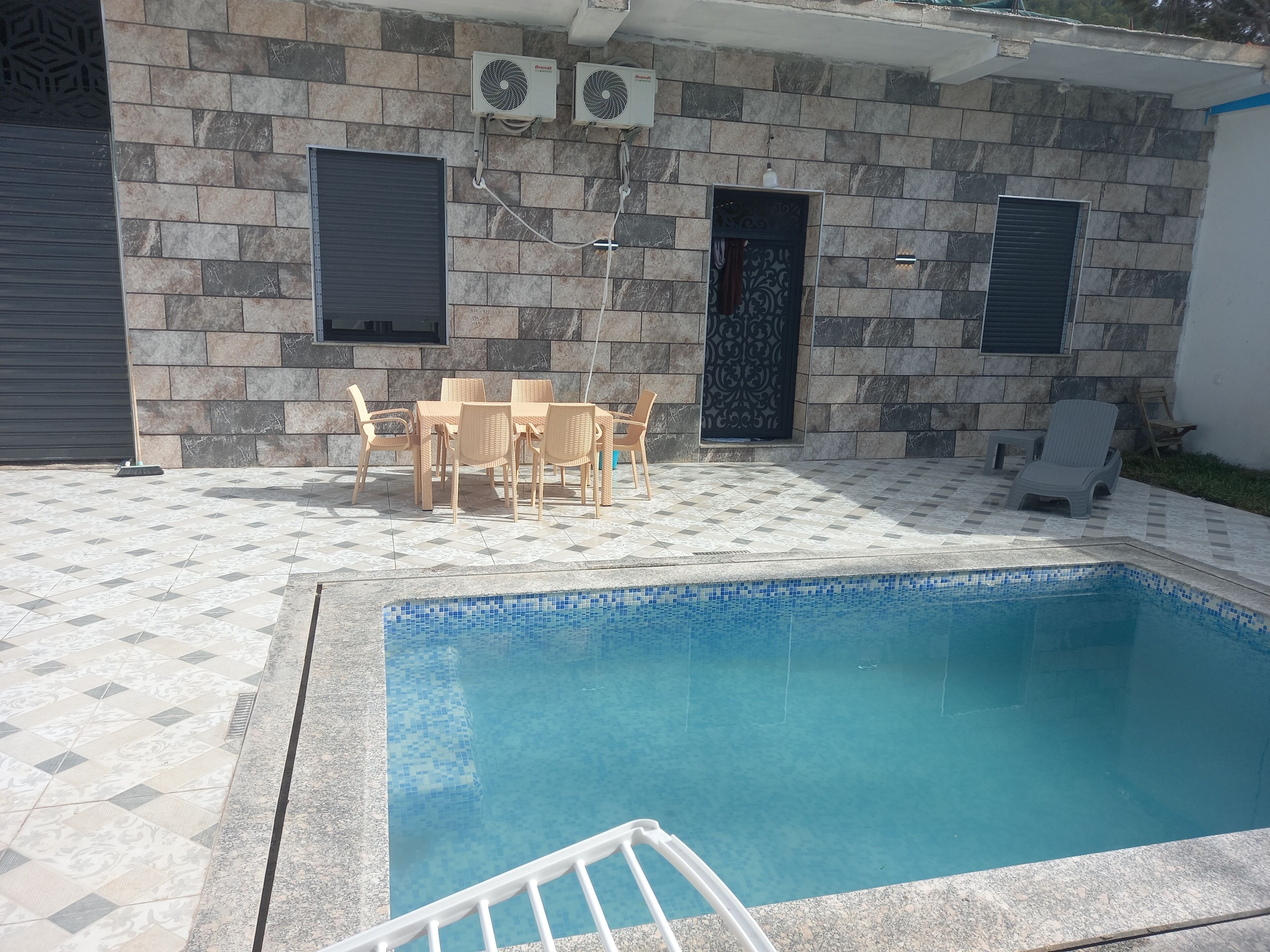 Une piscine avec une terrasse carrelée, une table avec des chaises et deux unités de climatisation fixées au mur.