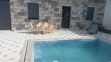 Une piscine avec une terrasse carrelée, une table avec des chaises et deux unités de climatisation fixées au mur.