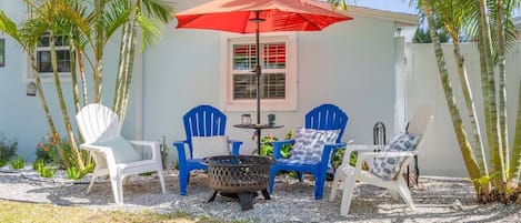 An outdoor seating area with two white and two blue Adirondack chairs, a red umbrella, and a fire pit.
