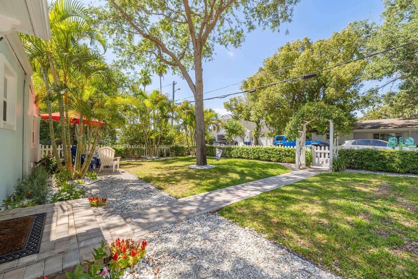 A residential area with a paved pathway, a white picket fence, and a variety of trees and shrubs.