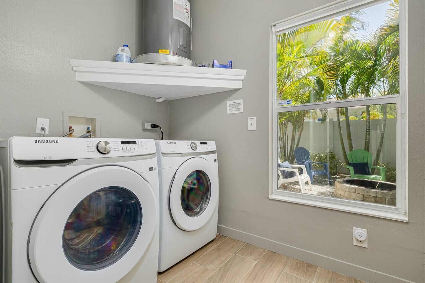 A laundry area with two Samsung washing machines, a white shelf above, and a window with a view of palm trees.