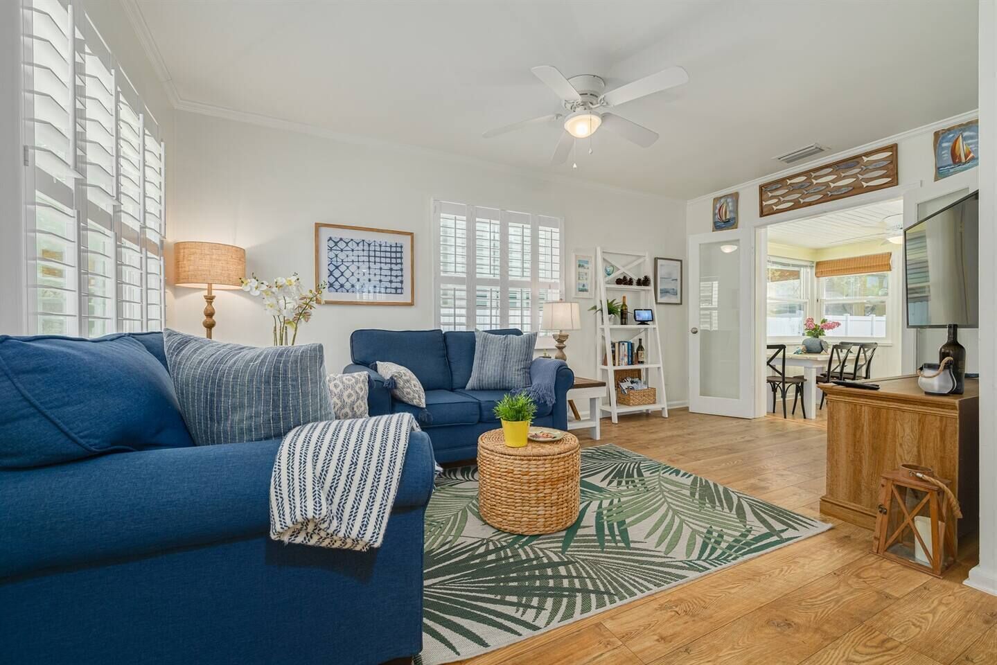 A living room with a blue sofa, a coffee table, and a patterned rug. There is a bookshelf and a dining area visible in the background.