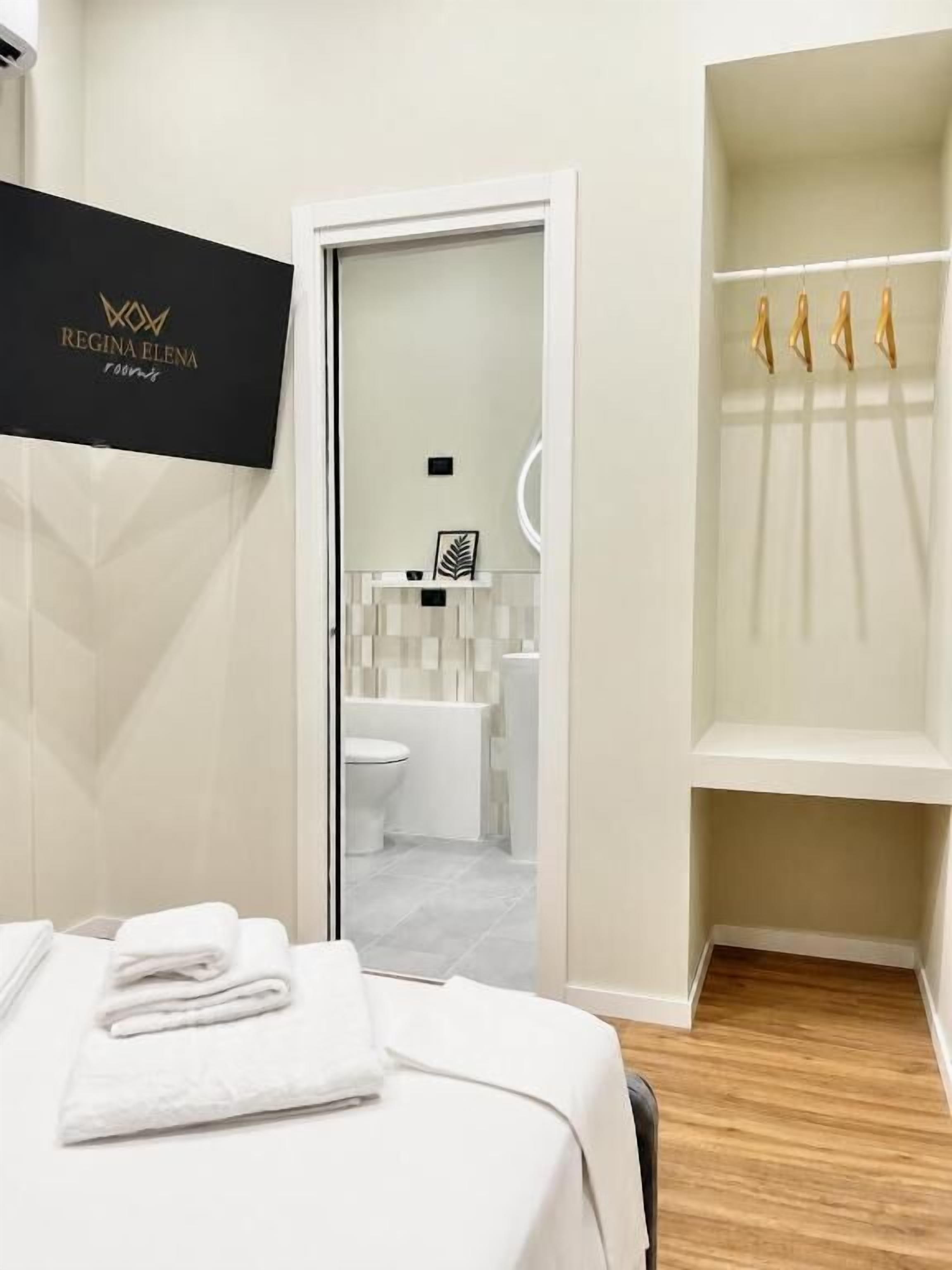 A hotel room with a bed, a white wall, a door leading to a bathroom, and a shelf with towels.