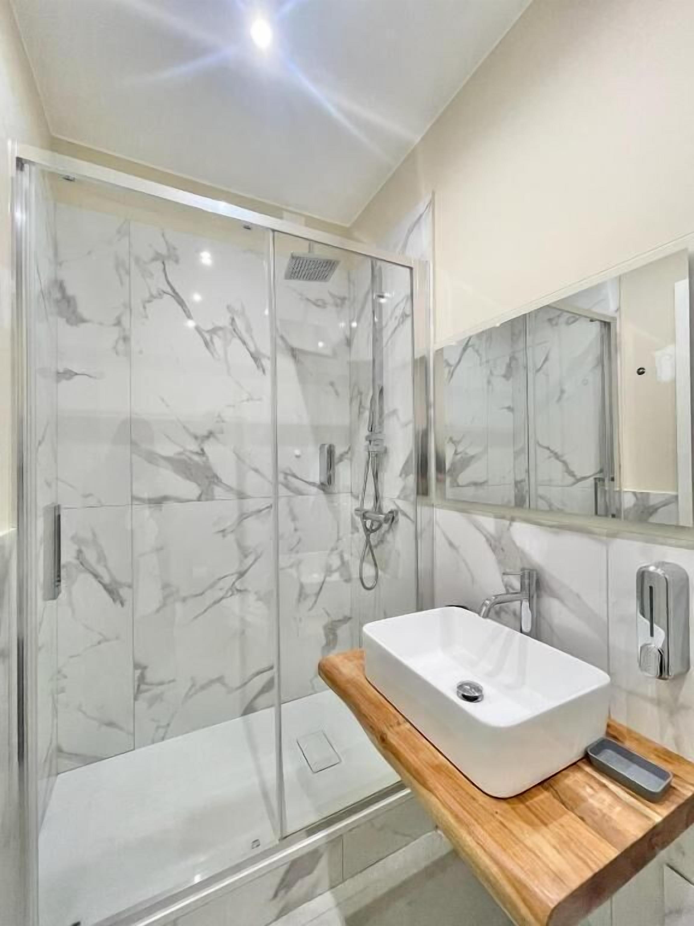 Un bany modern amb plat de dutxa de vidre, un lavabo blanc i una superfície de taula de fusta.