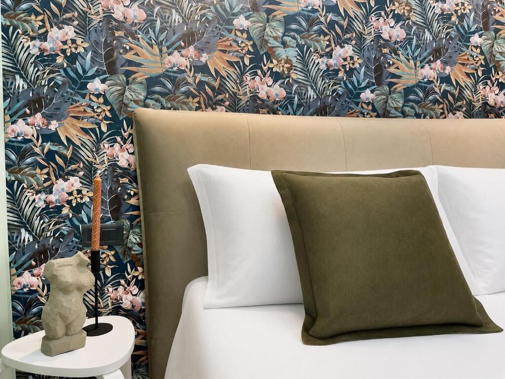 Een bed met een beige hoofdeinde, witte en olijfgroene kussens en een behang met bloemen.