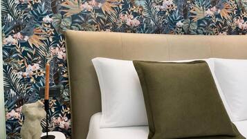Een bed met een beige hoofdeinde, witte en olijfgroene kussens en een behang met bloemen.