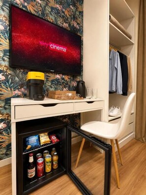 Habitación de hotel compacta con televisor de pantalla plana en la pared, una kitchenette pequeña con cafetera, un mini refrigerador con bebidas y una silla blanca.