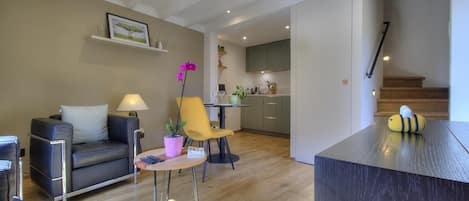 Una sala moderna con un sofá, una mesita y una silla. Se observa una cocina al fondo con armarios y una encimera.