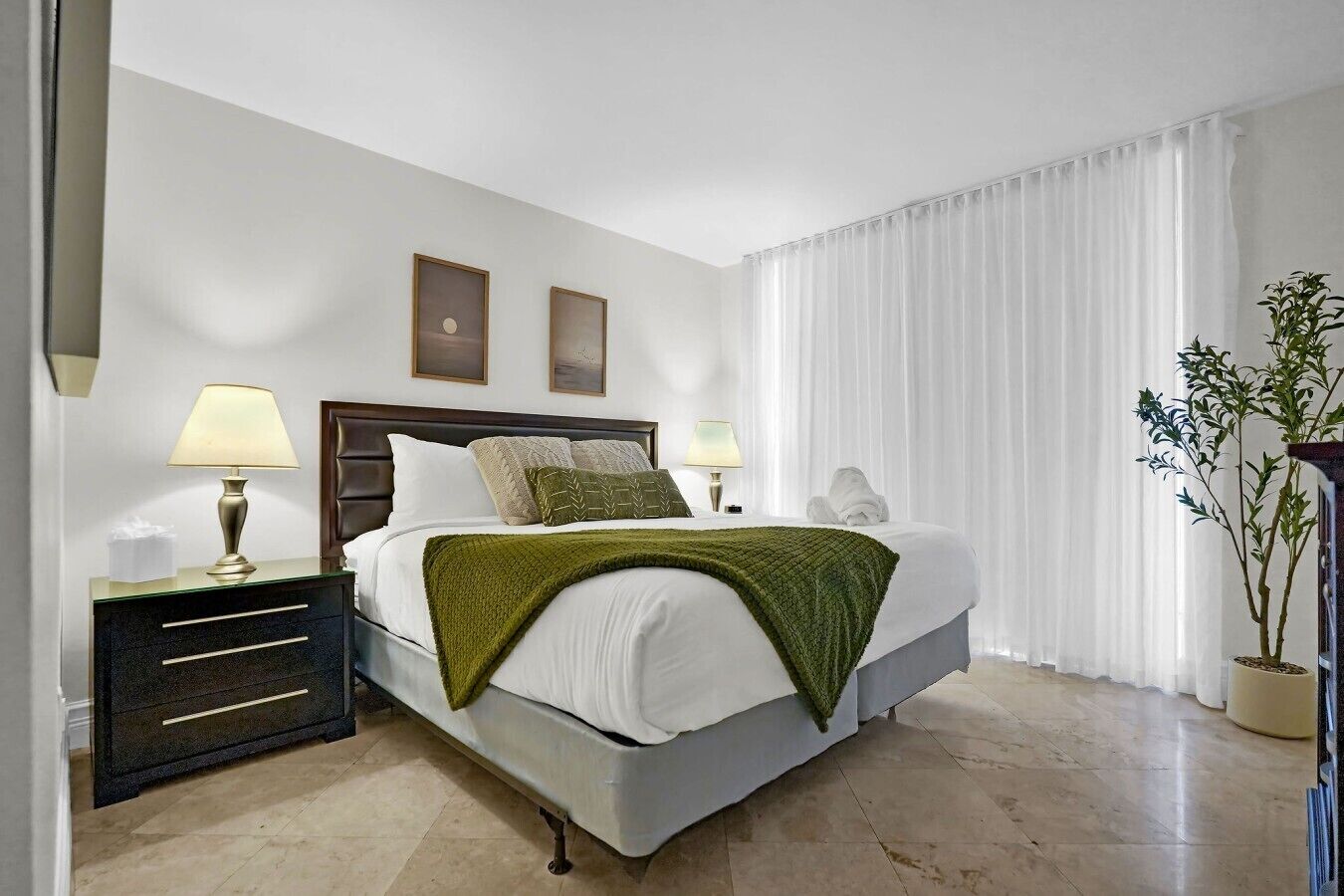 Hotelkamer met een bed, twee bedlampjes, een nachtkastje, een spiegel, een raam met witte gordijnen en een bloempot met plant.