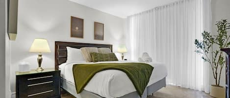 A hotel room with a bed, two bedside lamps, a nightstand, a mirror, a window with white curtains, and a potted plant.