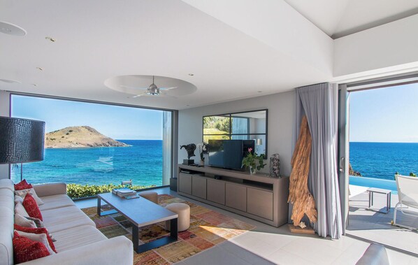 A modern living room with a large window overlooking the sea, a flat-screen TV, and a dining area with a view.
