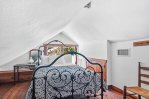 A bedroom with a sloped ceiling, a bed with ornate metal headboard, a wooden chair, and a small table with a lamp. - 1844 Alsatian Home on the Medina River (Castroville)