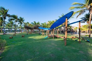 Un parque infantil con columpios y un tobogán, rodeado de palmeras y césped.