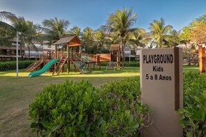 Zona de juegos con tobogán y estructuras para escalar, rodeada de palmeras y follaje.