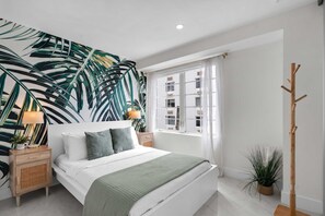 Un dormitorio con cama, mesita de noche, lámpara, ventana y perchero.