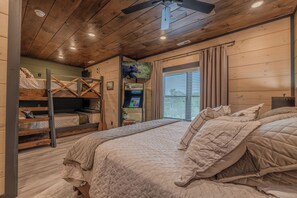 Ein schlicht eingerichtetes Schlafzimmer im Blockhausstil mit Etagenbett, Flachbildfernseher, kleinem Tisch und einem Fenster mit Jalousien.