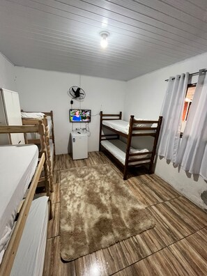 Una habitación con literas, un escritorio pequeño con un ventilador, una televisión y una cortina blanca.