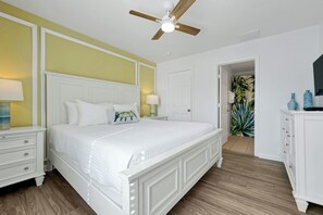 Ein Schlafzimmer mit einem weißen Bett, einem Deckenventilator, einem Nachttisch mit Lampe und einer Tür zu einem anderen Raum.