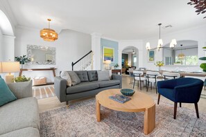 Ein modernes Wohnzimmer mit einem grauen Sofa, einem runden Couchtisch und einem blauen Sessel. Es gibt einen Essbereich mit Tisch und Stühlen, und im Hintergrund ist eine Küche zu sehen.