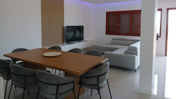 A modern living room with a wooden dining table, grey chairs, a grey sofa, and a television on a white cabinet.